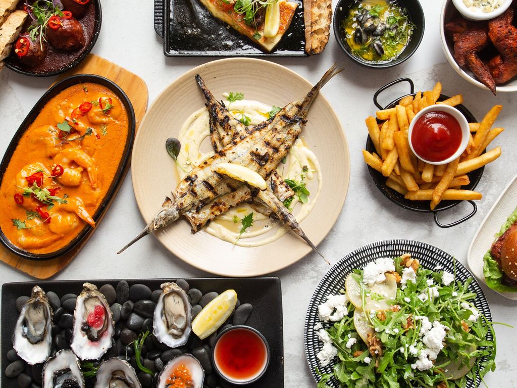 Une tablée vue de dessus : gambas à l'espagnole, poisson grillé, huîtres sur galets, salade de chèvre et frites.